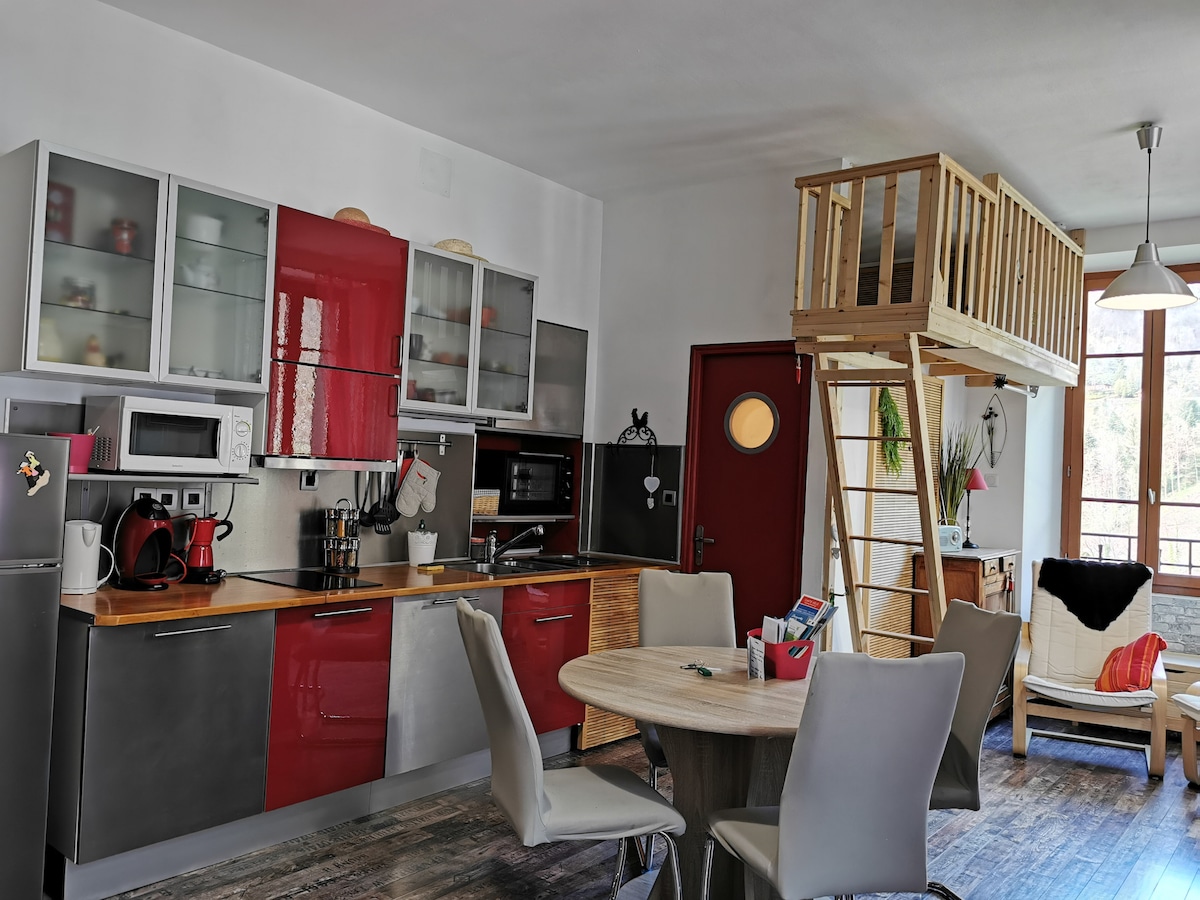 The kitchen area is equipped with modern appliances and colorful cabinetry, featuring a combination of gray and red finishes. A round wooden dining table is surrounded by four neutral chairs. A lofted sleeping area is visible with a wooden railing, adding vertical living space.