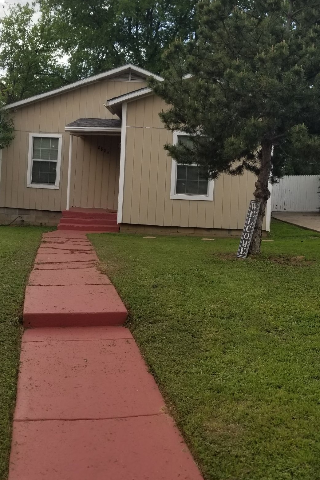 A well-maintained exterior of a single-story house is observed, featuring light-colored siding and a welcoming front porch. A neatly trimmed yard with green grass surrounds a red pathway leading to the entrance. A 'Welcome' sign is positioned beside a small pine tree.