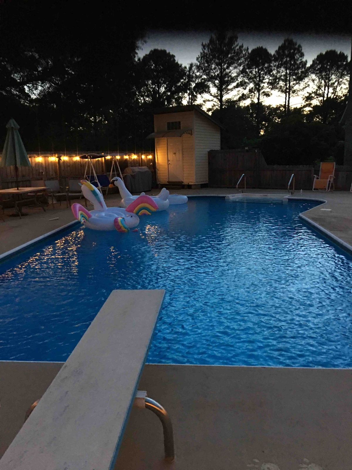 A spacious pool area is highlighted, featuring a vibrant blue pool surrounded by a concrete deck. Two large inflatable unicorns float serenely on the water's surface. Soft lighting from hanging bulbs illuminates the space, and tall trees line the background, creating a tranquil environment.