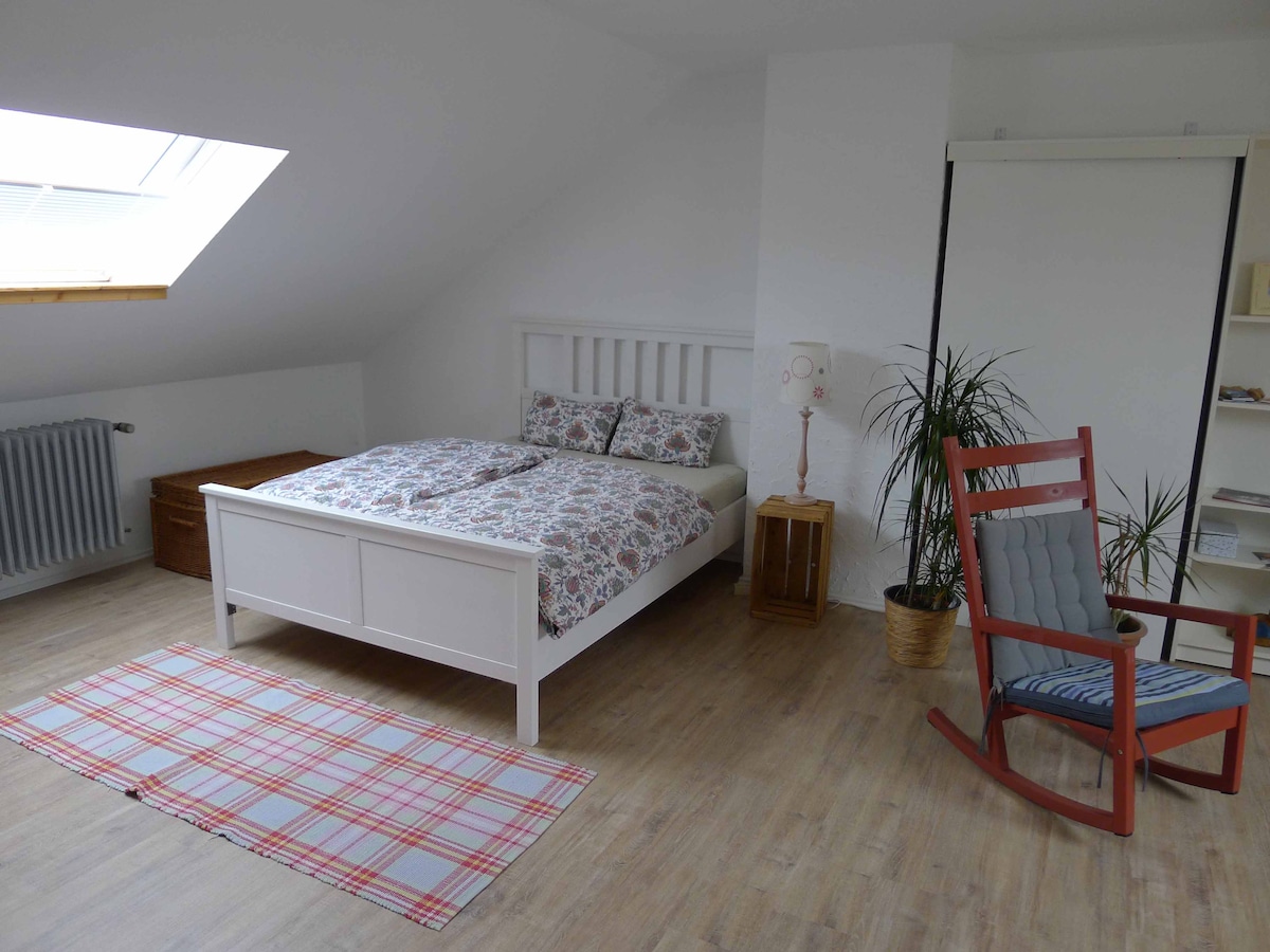 A spacious room features a white bed with a floral duvet and two pillows. A rocking chair is positioned beside a potted plant and a light source. Storage options include a wooden chest and shelves. Natural light enters through a skylight, enhancing the bright atmosphere.