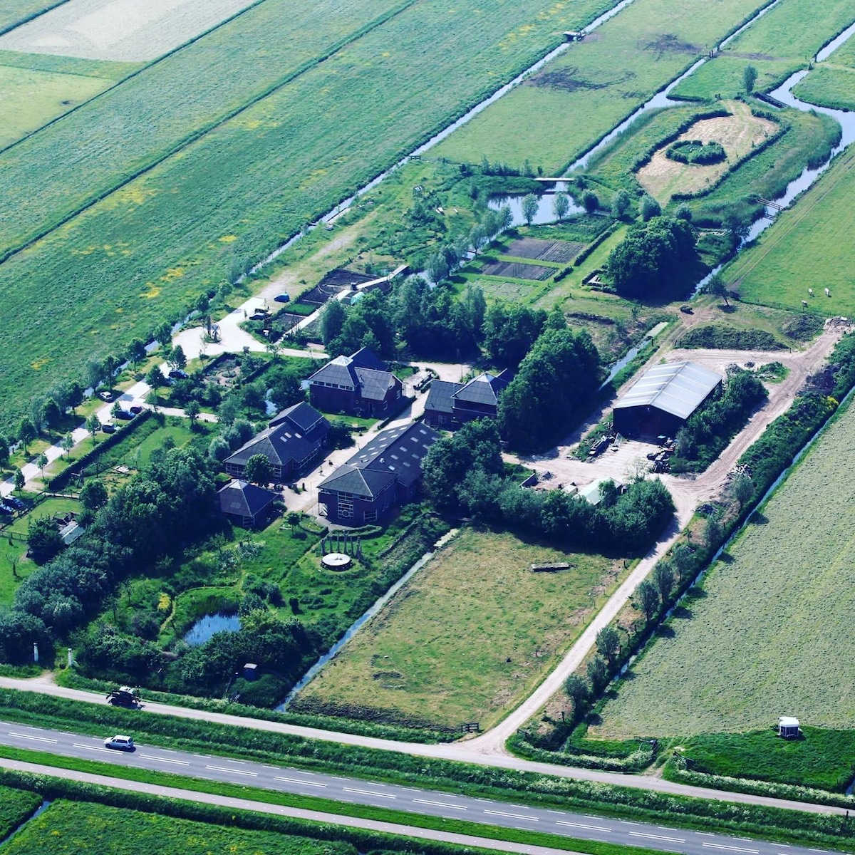 An aerial view of a farm landscape showcases the Eemlandhoeve surrounded by expansive fields. Various agricultural structures are visible, along with patches of gardens and water features. Roads connect different areas of the property, emphasizing the integration of nature and farming.