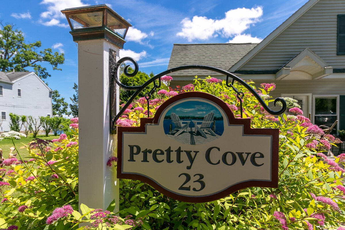A welcoming sign displays the name 'Pretty Cove 23', framed by lush green foliage and vibrant pink flowering plants. The clear blue sky serves as a contrasting backdrop, enhancing the inviting appearance of the entryway to the property.