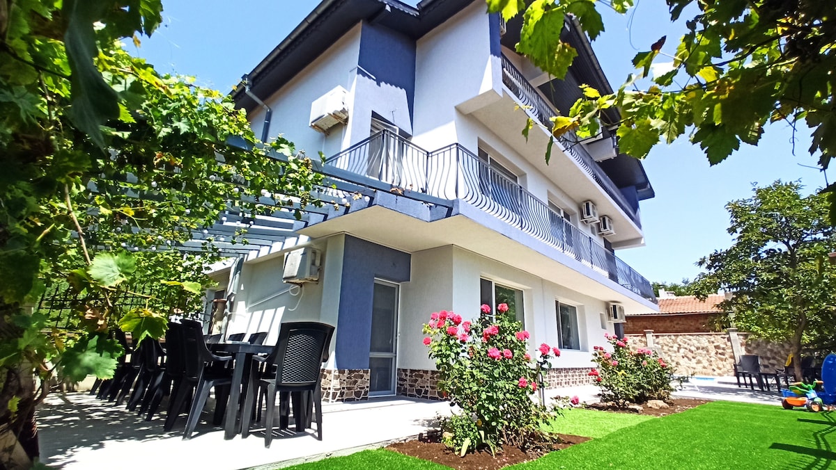 The villa showcases a multi-level structure with a modern exterior. A patio area features black chairs and tables, surrounded by blooming pink roses and a lush green lawn. Vines cover part of the balcony, creating a serene outdoor setting.