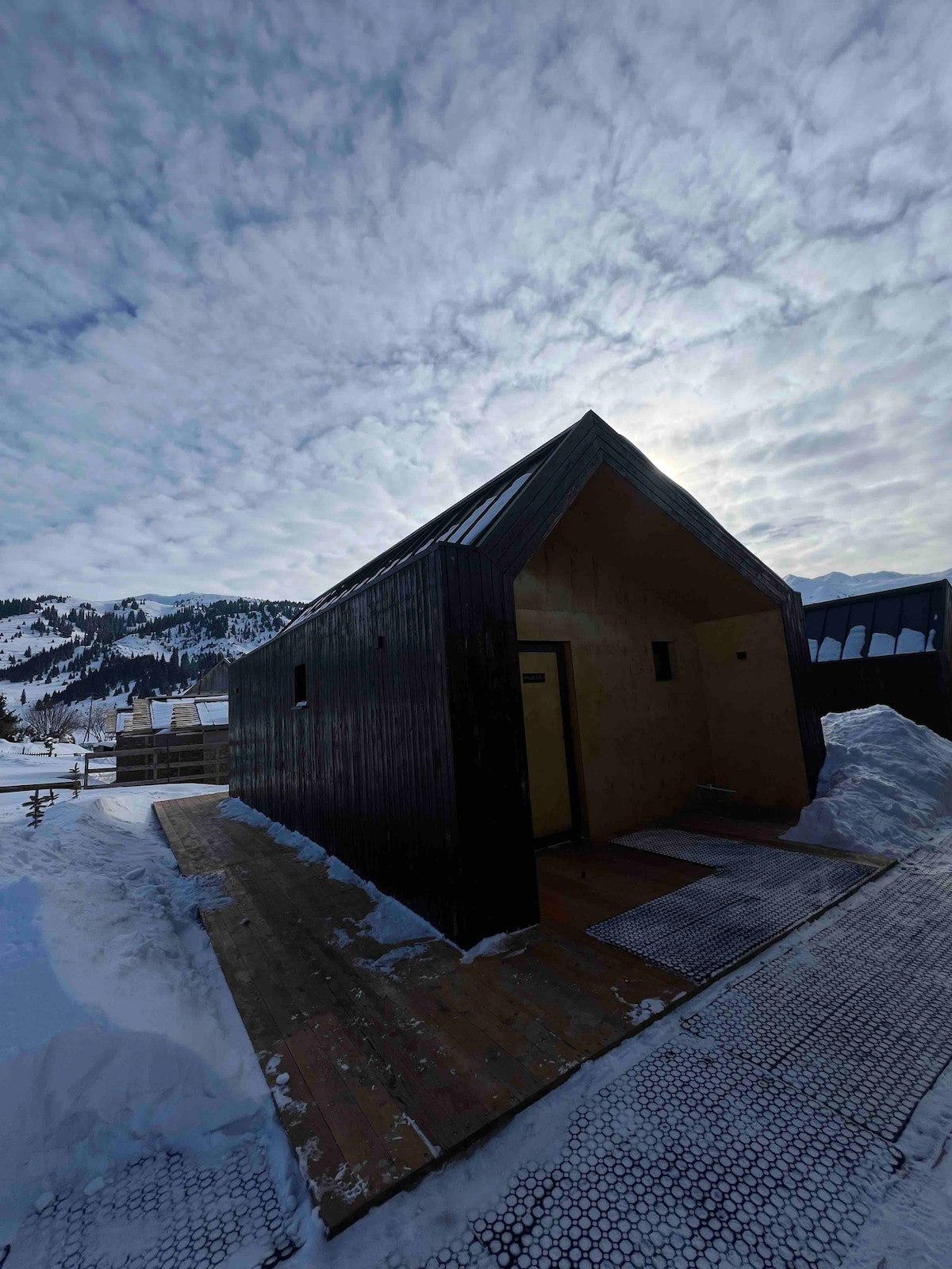 A modern cottage features a steeply pitched roof and dark exterior, set against a backdrop of snow-covered mountains. Wooden decking extends in front, leading to a main entrance. The sky is partially cloudy, casting soft light over the surroundings.
