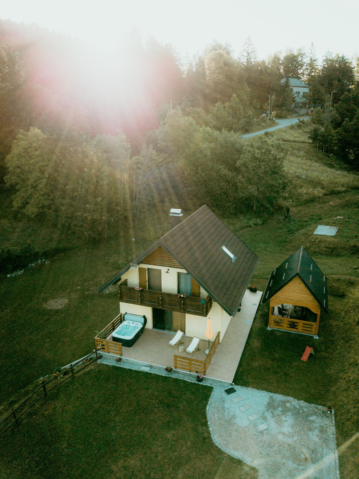 An aerial view of the holiday house 'Mavis', showcasing its rustic architecture surrounded by trees. A patio area features outdoor seating and a hot tub, while a small shed is visible nearby. Sunlight gently illuminates the scene, enhancing the natural landscape.
