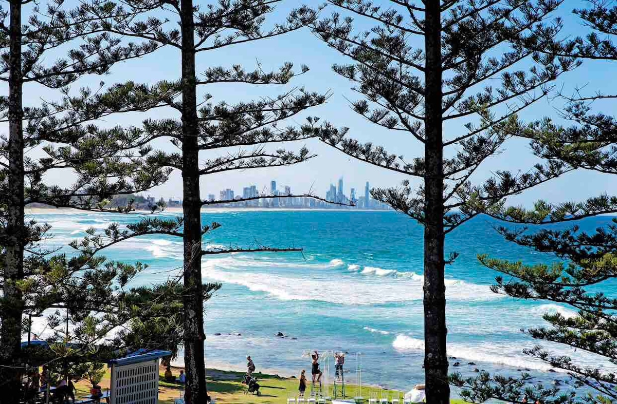 The image captures a scenic view of the ocean framed by tall pine trees. Gentle waves roll onto the shore, and distant city skyscrapers rise against a clear blue sky. People can be seen enjoying the beach area, adding to the lively atmosphere.