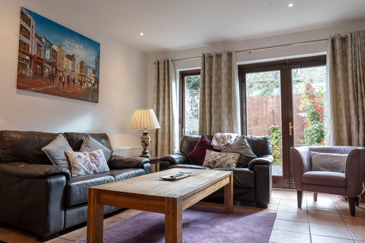 A living area displays two dark leather sofas adorned with decorative pillows, accompanied by a light wooden coffee table. Natural light enters through glass doors leading to a garden, while a piece of artwork depicting a street scene adds a touch of visual interest to the wall.