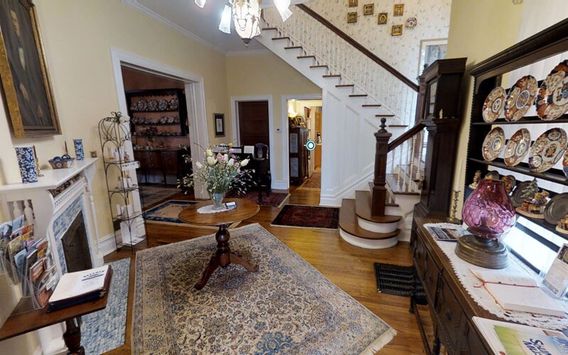 An inviting foyer features a round wooden table centered on a floral area rug. A staircase with wooden handrails leads to the upper level. Display cabinets with decorative plates and a lamp add character, while floral arrangements and brochures sit on a nearby side table.