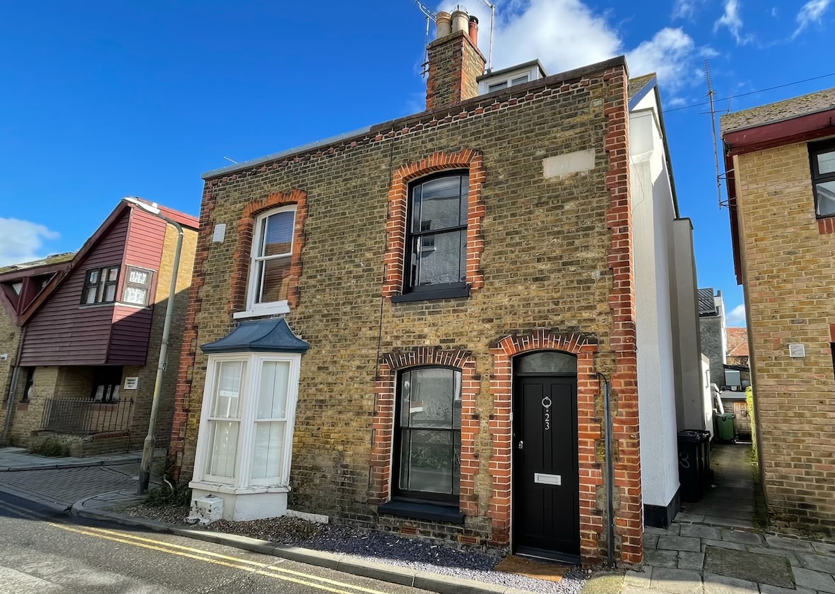 The cottage exterior presents a mix of historic and modern design, featuring a mixture of brick and painted surfaces. A distinctive bay window is located on the left, while the main entrance is positioned centrally. Clear blue skies serve as a backdrop, enhancing the inviting atmosphere.