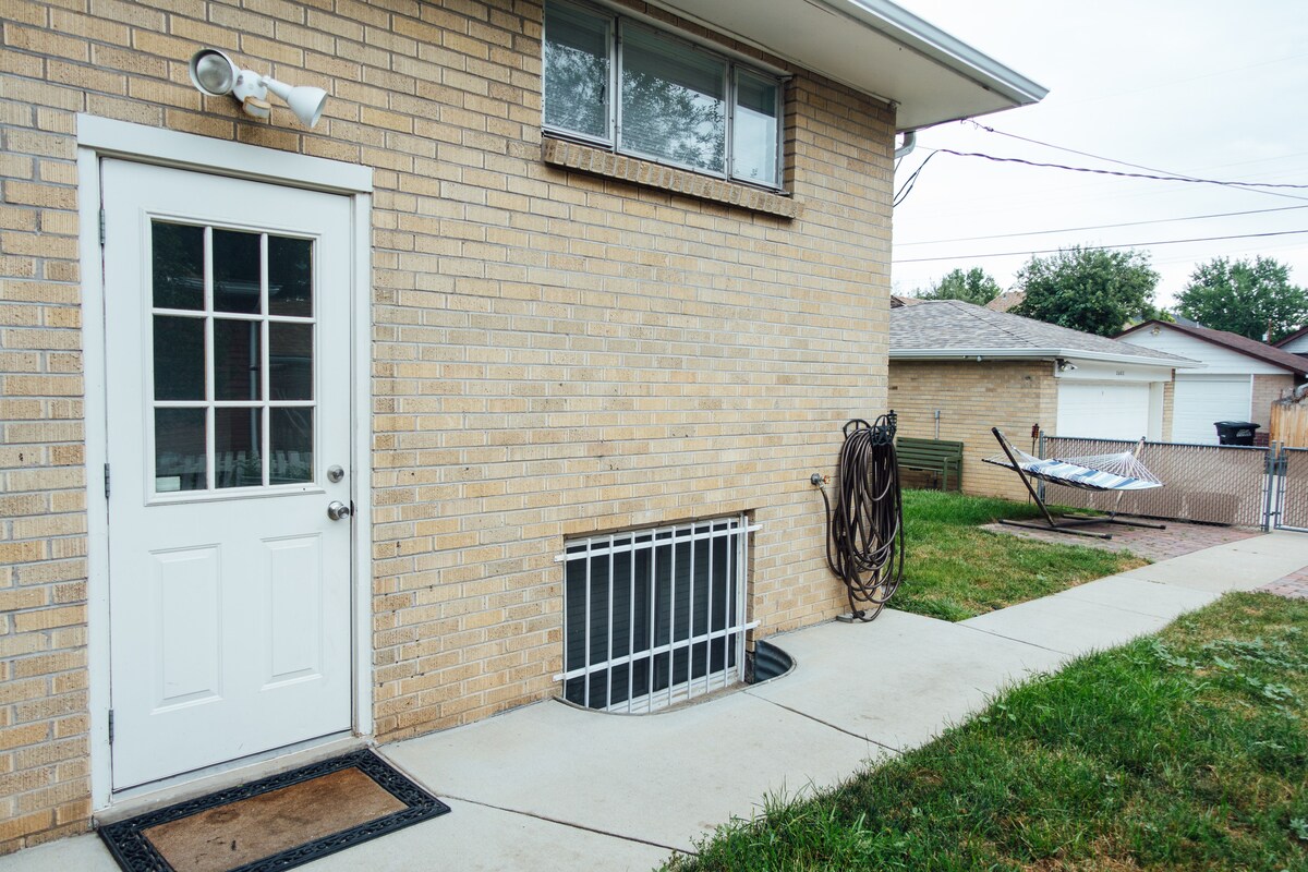 A private entrance to a garden-level apartment is visible, featuring a white door with glass panels. Adjacent to the entrance is a small window well covered with a security grate. A neatly paved walkway leads to the outdoor space, showing adjacent grassy areas and a hammock.