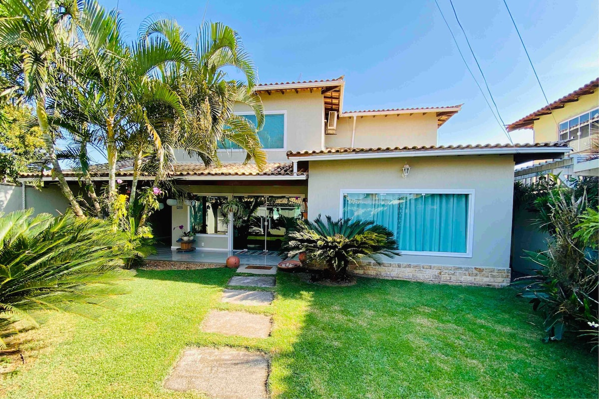 The exterior of a spacious house is framed by lush greenery and palm trees, with a well-maintained garden. A stone pathway leads to the entrance, which features large windows letting in natural light. The blue sky enhances the inviting atmosphere of the home.