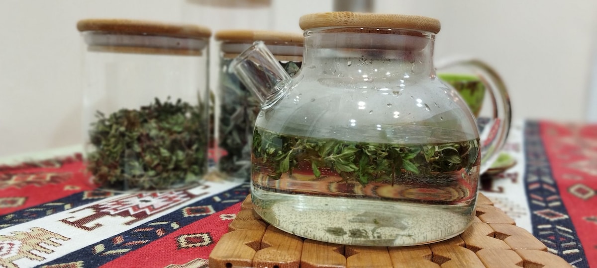 A glass teapot filled with brewed herbs is positioned on a wooden mat. Behind it, three jars containing dried herbs are visible. The teapot's clear contents display the vibrant green of the steeping leaves, resting on a patterned tablecloth with rich colors.