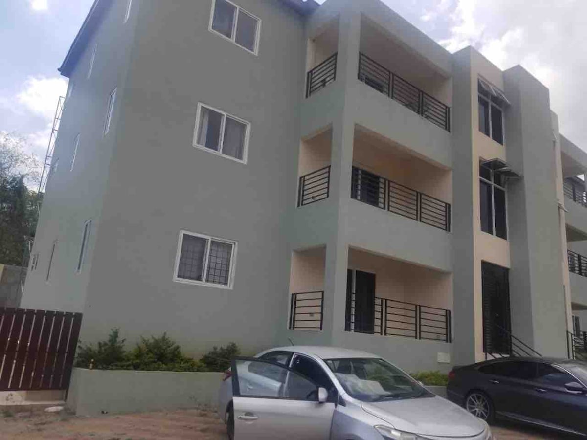 A three-story building features a modern design with a light green exterior. Multiple balconies are visible, showcasing black railings. Two vehicles are parked in the foreground, and manicured shrubs line the base of the structure.