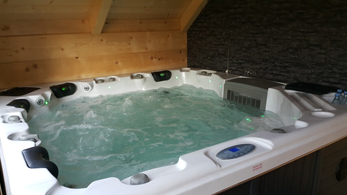 A spacious hot tub is positioned indoors, featuring multiple seating areas with jets for relaxation. Soft lighting creates a calming atmosphere, while the water appears inviting and warm. Natural wood accents on the ceiling and stone walls add to the soothing environment.