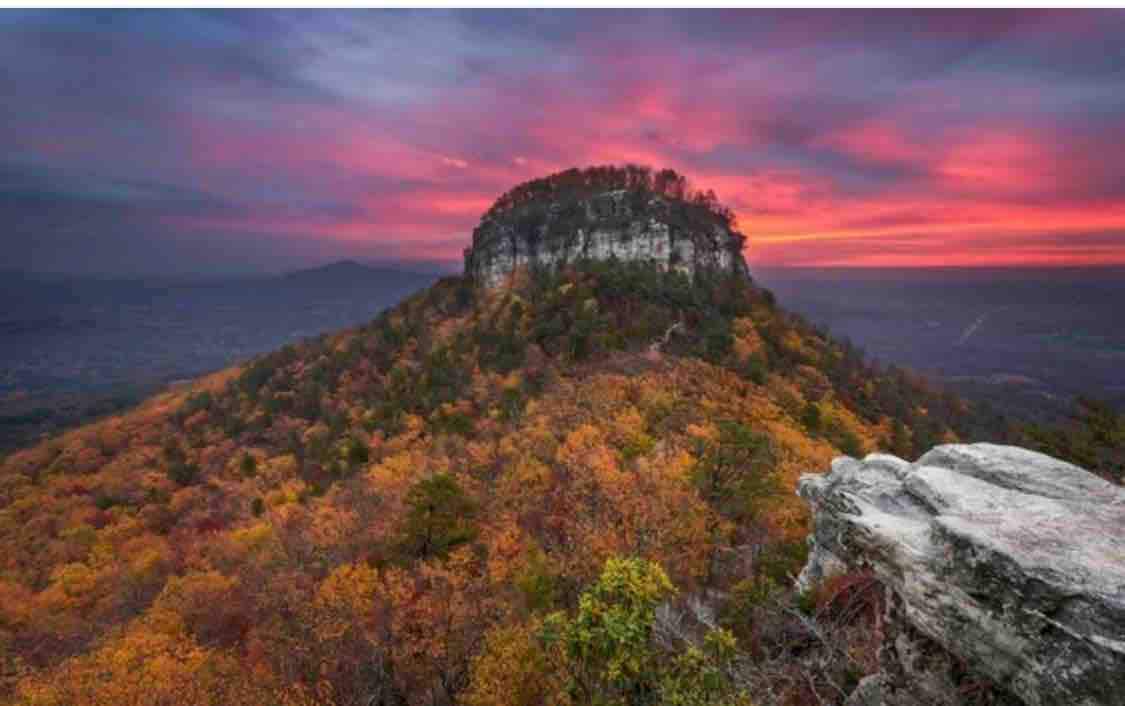 The image captures a mountain landscape during sunset, with vibrant colors illuminating the sky above a prominent rocky peak. Surrounding trees showcase autumn foliage, transitioning into shades of gold and orange, while rugged rock formations frame the foreground.