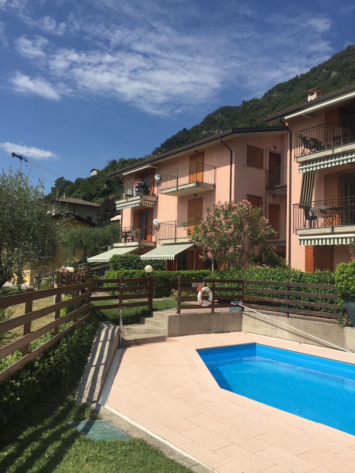 A well-maintained outdoor area features a pool surrounded by neat walkways and landscaped gardens. The bright facades of the nearby buildings are complemented by greenery and flowering plants, while blue sky and mountains create a serene backdrop.