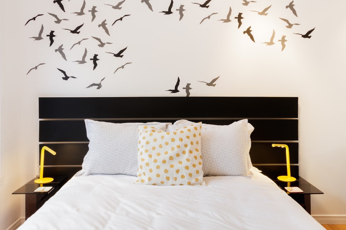 A spacious bed is positioned against a white wall, adorned with a striking silhouette of flying birds. Two small yellow lamps are placed on either side of the bed, and decorative pillows with a polka dot pattern add a touch of comfort.