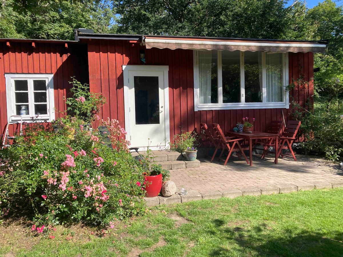 A charming red wooden exterior with white-framed windows is showcased. A stone-paved patio area features a dining table with chairs, surrounded by vibrant flowering plants and greenery. The entrance door is centrally located, inviting guests to the cozy space inside.