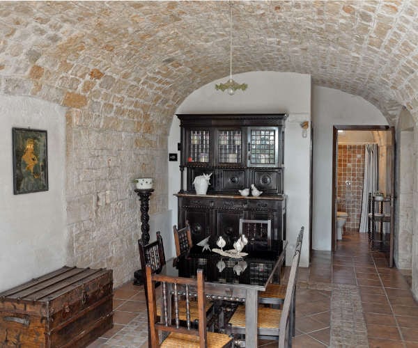 A rustic dining area features a dark wooden table surrounded by six chairs. An antique sideboard with glass paneling showcases decorative items. The vaulted stone ceiling adds character, while a doorway leads to a modern bathroom space visible in the background.