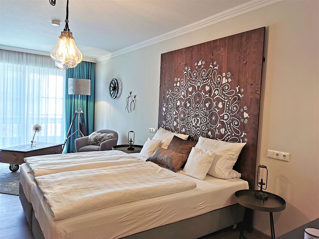 A spacious double bedroom features a large headboard with intricate designs above two neatly made beds. Soft white linens complement the neutral tones of the decor. A cozy seating area with a chair is positioned near a large window that allows natural light to illuminate the space.