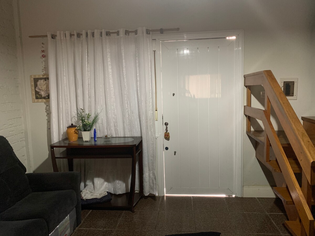 The entrance area features a white front door with a small window, complemented by light-colored curtains that frame the space. A dark wooden table is positioned nearby, displaying a small plant and decorative items. Stairs leading to the upper level are visible on the right.