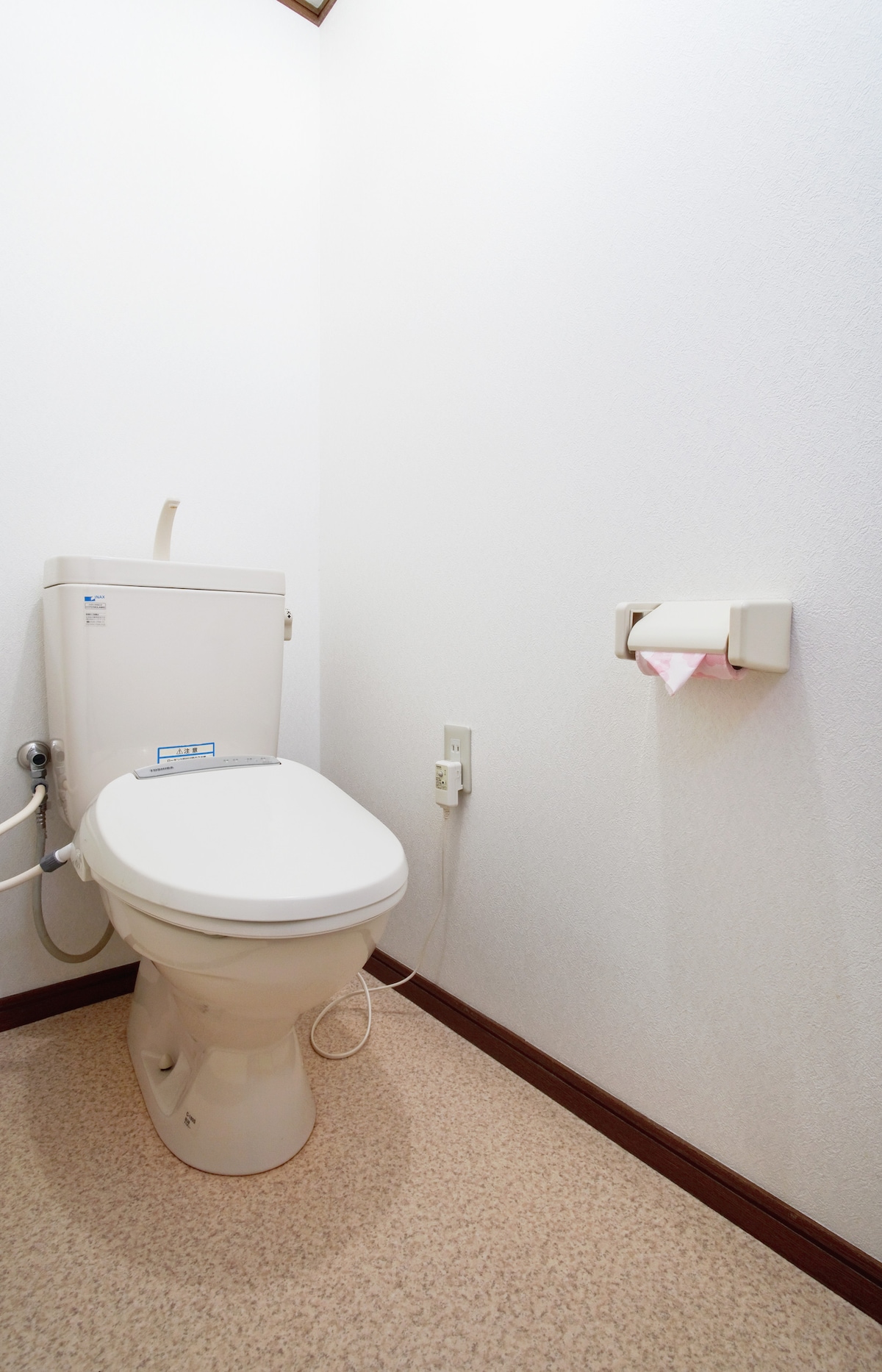 A clean and simple restroom is presented, featuring a modern toilet and a discrete toilet paper holder. The walls are plain and the flooring is light-colored, providing a minimalist and tidy appearance.