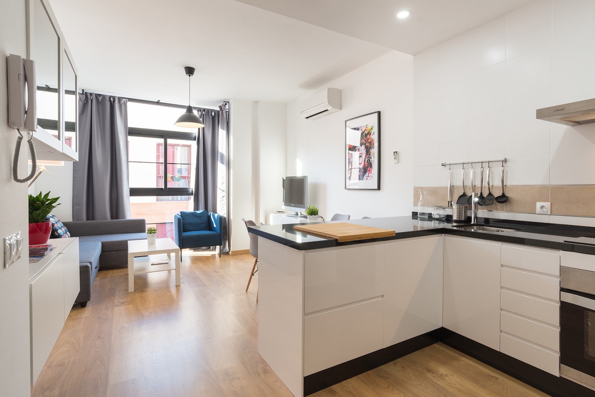 An open-concept living area is shown, featuring a well-equipped kitchen with a wooden countertop and stylish cabinetry. A comfortable seating area includes a gray sofa and a small coffee table, with natural light streaming through large windows draped with gray curtains.