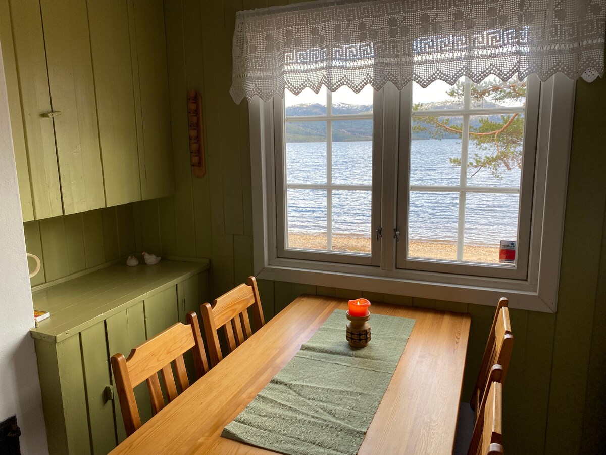 A cozy dining area is set with a wooden table and chairs, complemented by a simple table runner. Natural light filters through a large window adorned with lace curtains, offering a view of the water and surrounding landscape.