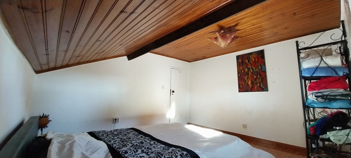 A simple and cozy sleeping area is presented, featuring a spacious bed dressed in white linens and a patterned blanket. The wooden ceiling adds warmth, while a colorful wall art piece provides a pop of color. Natural light filters in from a nearby door.