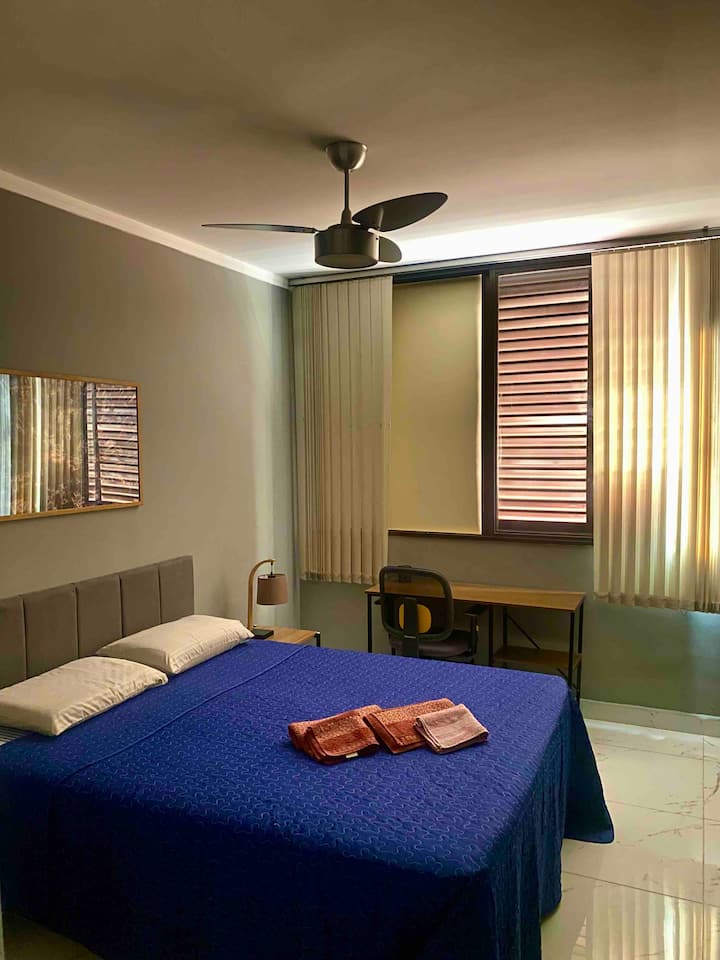 Queensize bed, bureau, Flexform-stoel, 10.000 btu draagbare airconditioner en plafondventilator.