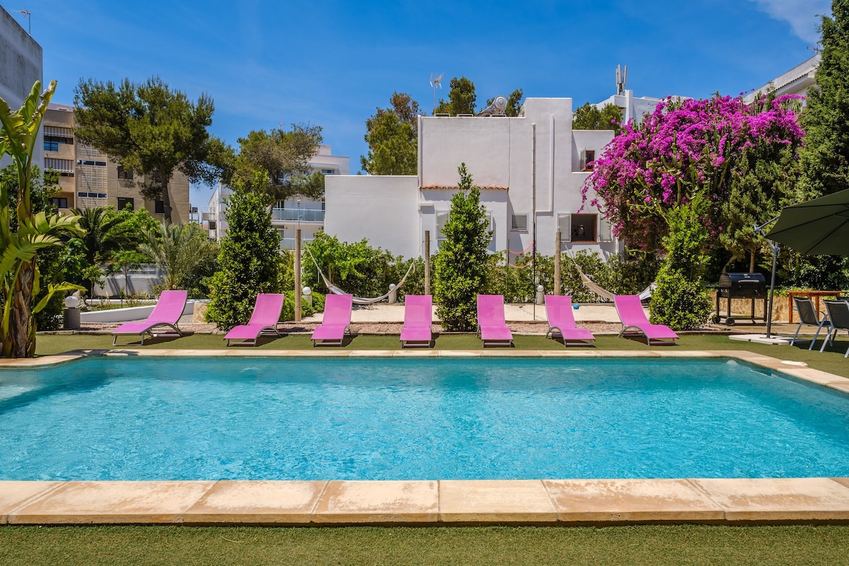 A tranquil outdoor area features a spacious saltwater pool surrounded by vibrant purple loungers. Lush greenery and blooming plants frame the pool area, providing a serene setting for relaxation. A grill can be seen nearby, complementing the inviting outdoor space.