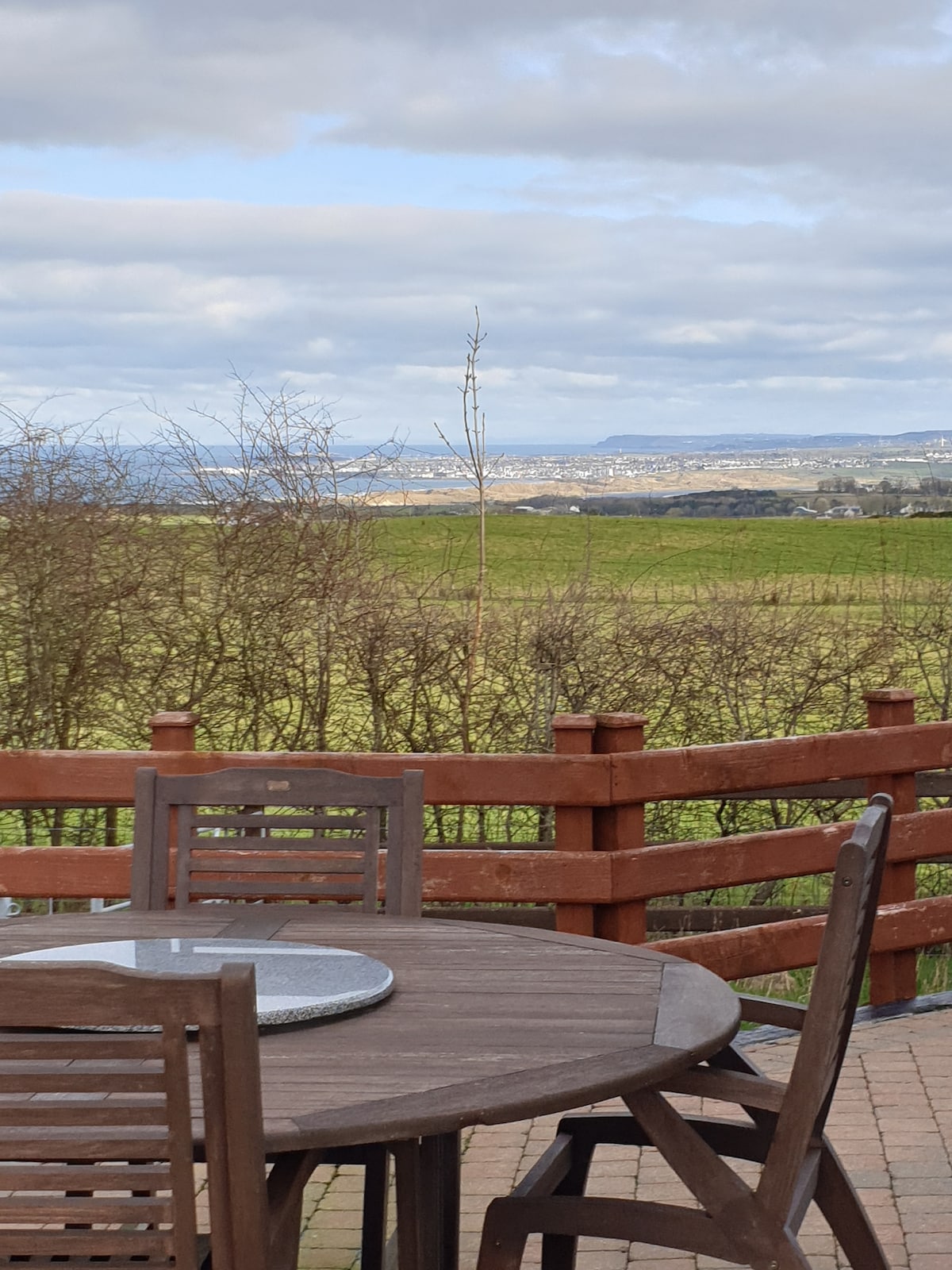A wooden table and chairs are arranged on a patio, providing a setting for outdoor dining. In the background, expansive fields stretch towards the horizon, framed by a wooden railing and a backdrop of distant hills under a cloudy sky.