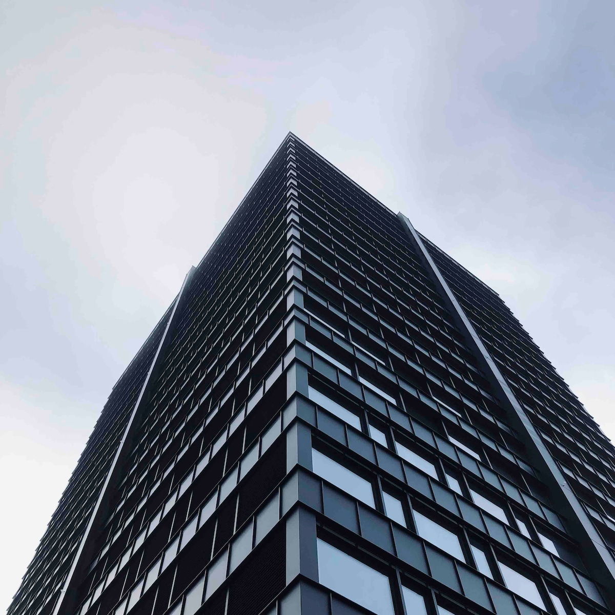 The image captures the towering facade of a modern high-rise building, featuring a grid of reflective glass windows that accentuate its sleek architectural design. The structure's sharp angles and clean lines are emphasized against a cloudy sky, conveying a sense of height and urban sophistication.