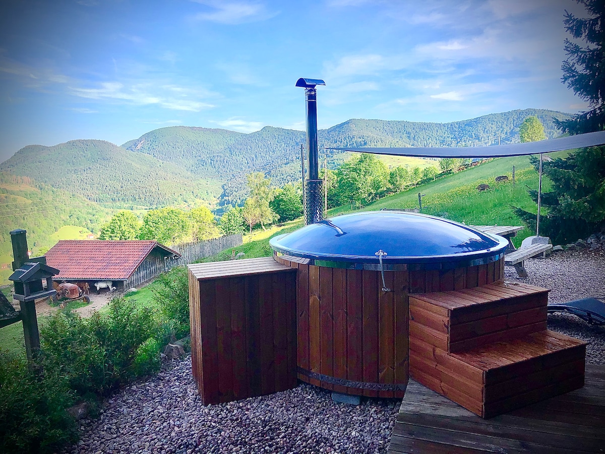 A nordic bath is positioned on a wooden deck, showcasing its circular wooden structure with a metallic chimney. The surrounding landscape features rolling green hills and mountains in the background, offering a serene view.
