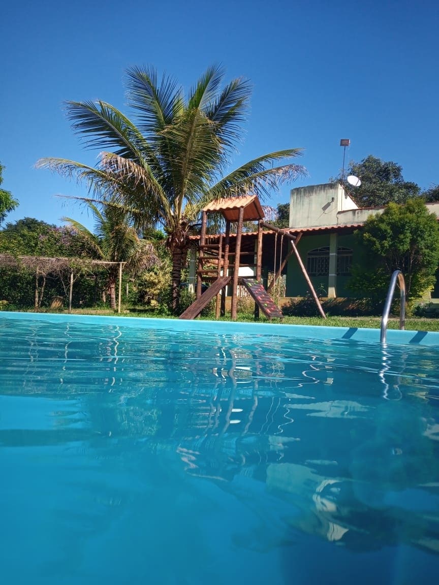 A tranquil scene featuring a clear blue pool with gentle ripples reflecting sunlight. A tall palm tree stands nearby, providing shade. In the background, a wooden play structure is visible, surrounded by greenery, contributing to a serene outdoor space.