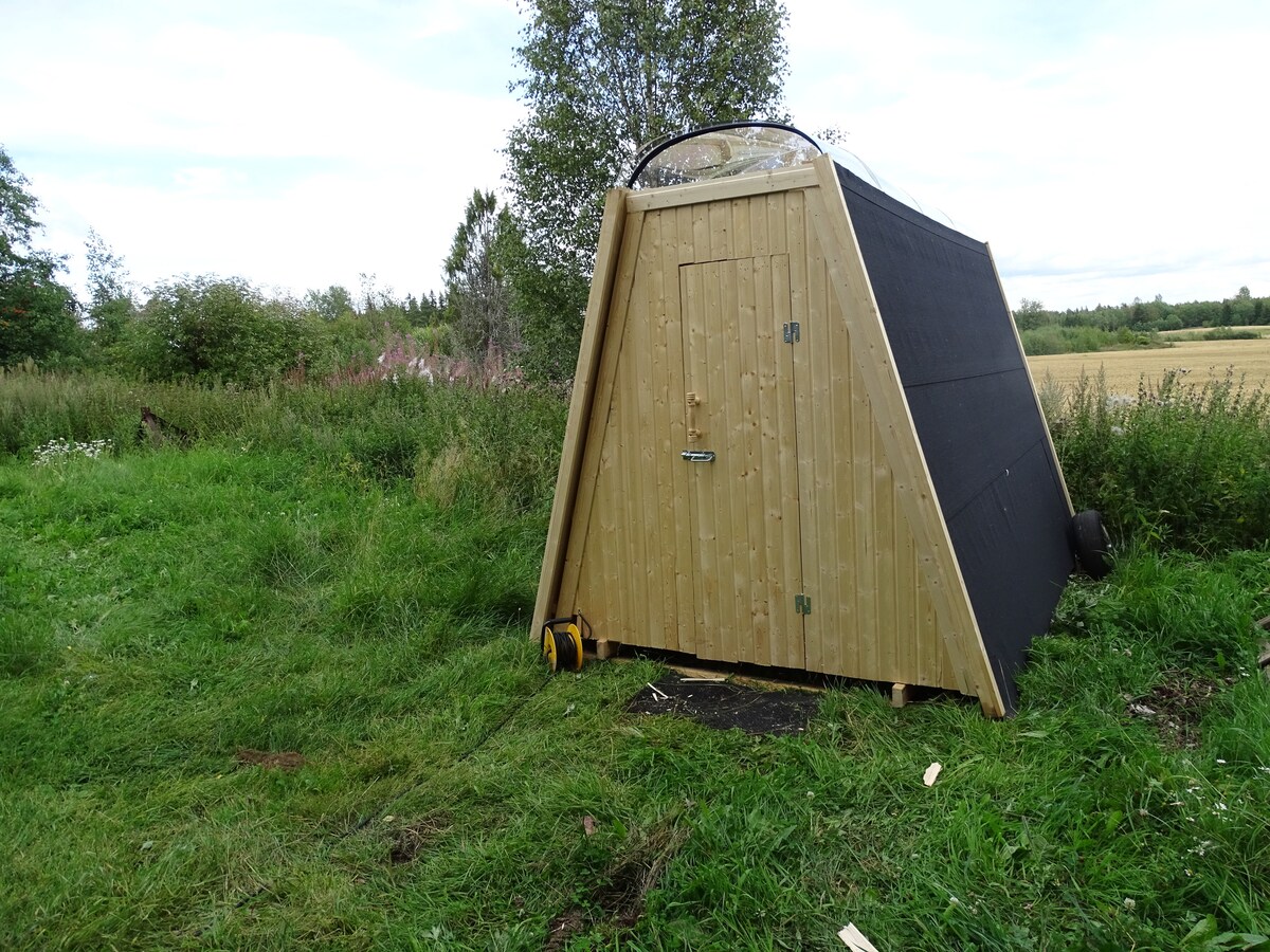 An A-frame cabin with wood paneling is set amidst lush green grass and wildflowers. Its large skylight dome is visible on top, allowing natural light to enter. The cabin is surrounded by open fields, with trees and shrubs in the background.