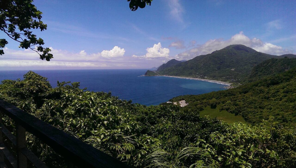 A panoramic view reveals a vast expanse of ocean meeting the lush green coastline, framed by mountains in the background. The sky is bright with scattered clouds, creating a serene and tranquil scene.