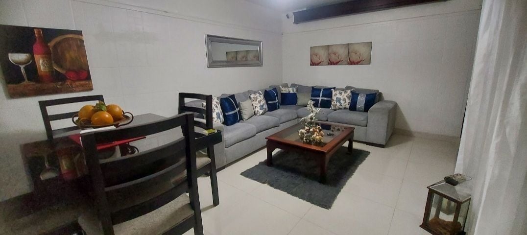 A spacious living area features a light-colored sectional sofa adorned with decorative cushions. A wooden coffee table sits atop a dark area rug, surrounded by artwork and a mirror on the walls. A dining table with chairs is visible, showcasing a centerpiece of fruits.