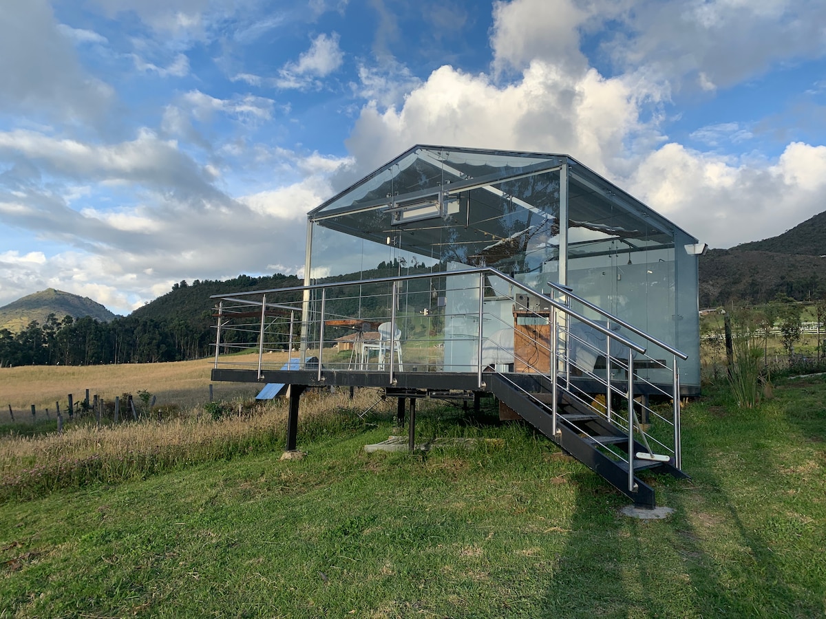A glamping accommodation with a modern glass structure sits elevated on a grassy area. A ramp leads to the entrance, providing accessibility. A scenic view of gently rolling hills can be seen in the background, framed by a mix of clouds and sunlight.