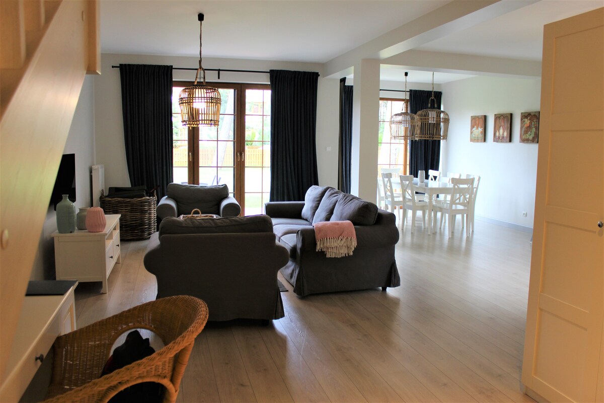 A spacious living area is visible, featuring two comfortable sofas and a coffee table. Large glass doors lead to an outdoor space. A dining area with a white table and chairs is located in the background. Light wood flooring complements the neutral-toned decor.