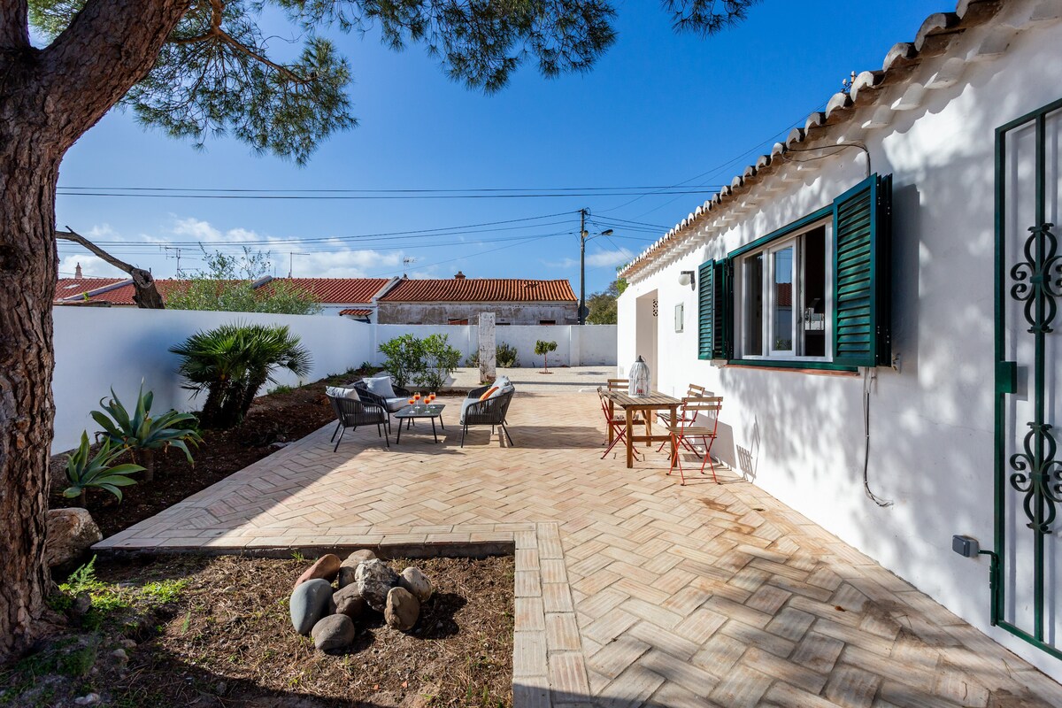 A spacious outdoor patio is highlighted by stone tiles and surrounded by greenery. Comfortable seating arrangements, including a couple of lounge chairs and a small dining table, offer a relaxing environment. Sunlight bathes the area, and the white cottage features green shutters and large windows.