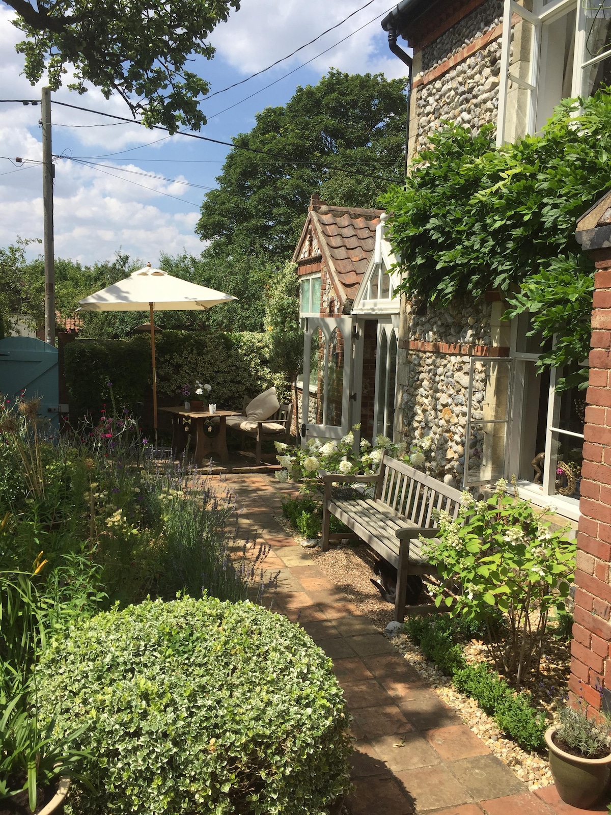 A garden space is presented, featuring a pathway lined with flowering plants and greenery. A wooden bench sits alongside a small table, and an umbrella provides shade. Stone walls, typical of flint construction, complement the vibrant natural setting.