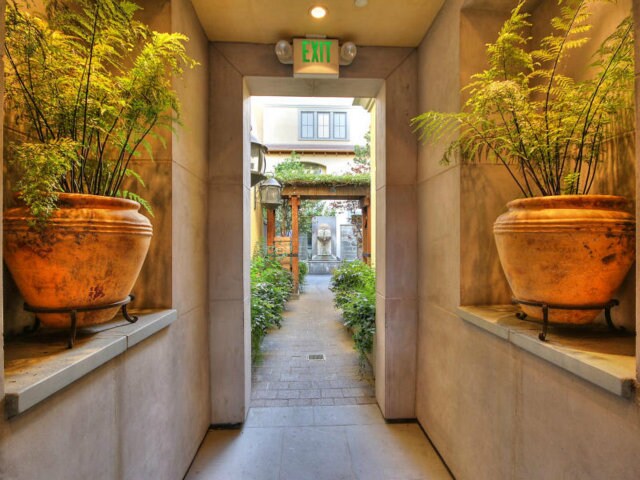 A well-lit corridor leads to an exit, framed by two large terracotta pots filled with lush greenery. Natural light filters in, creating a serene atmosphere. A glimpse of a garden path is visible beyond, enhancing the inviting appeal of the space.