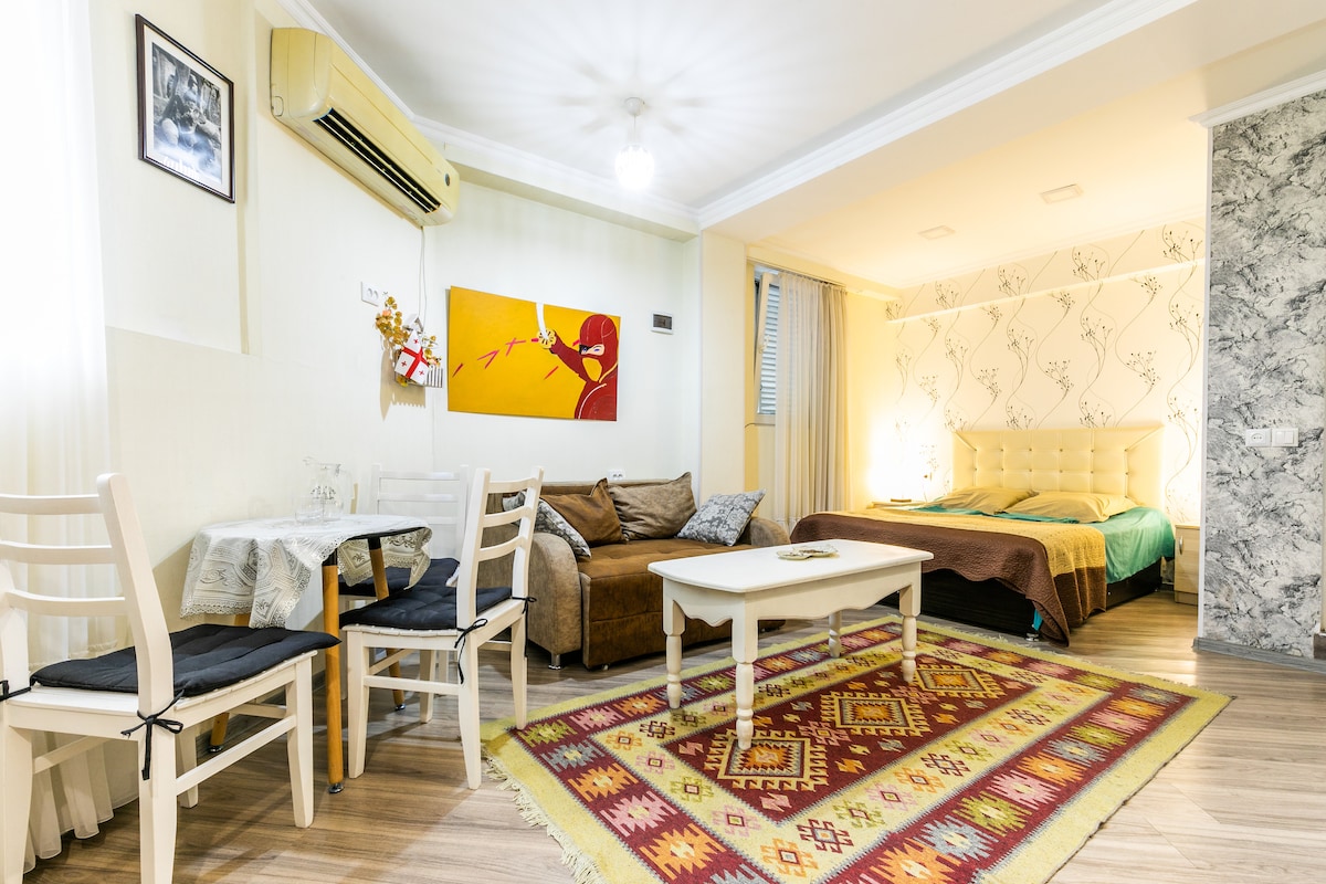 A studio flat is presented, featuring a spacious seating area with a brown sofa and a dining space with white chairs. Light-colored walls harmonize with patterned wallpaper. A cozy queen-sized bed is situated in the background, while an area rug adds warmth to the wooden flooring.