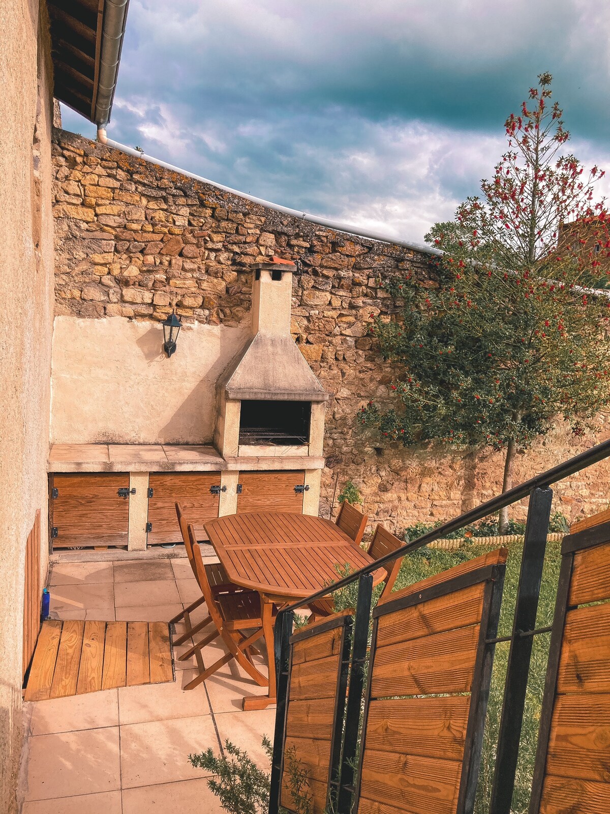 An outdoor terrace is presented, featuring a wooden dining table surrounded by matching chairs. A built-in stone barbecue is situated against a rustic stone wall, with greenery and a small tree adding a natural touch to the area.