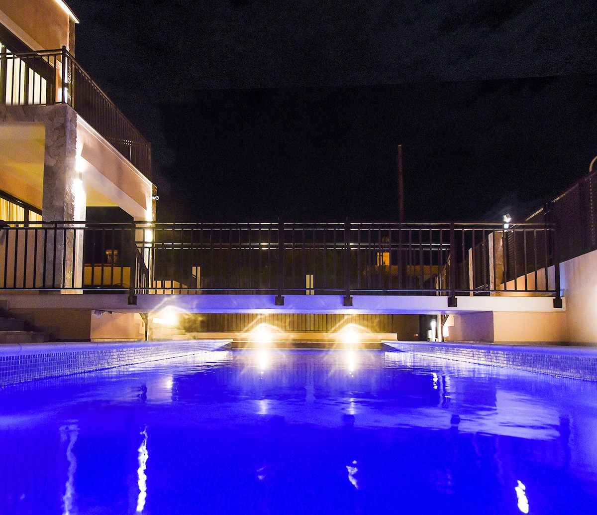 The heated swimming pool is illuminated with soft blue lighting, creating a serene night-time atmosphere. The pool's edge is flanked by the terrace and a railing, leading to a well-lit area of the patio and accessing the villa.
