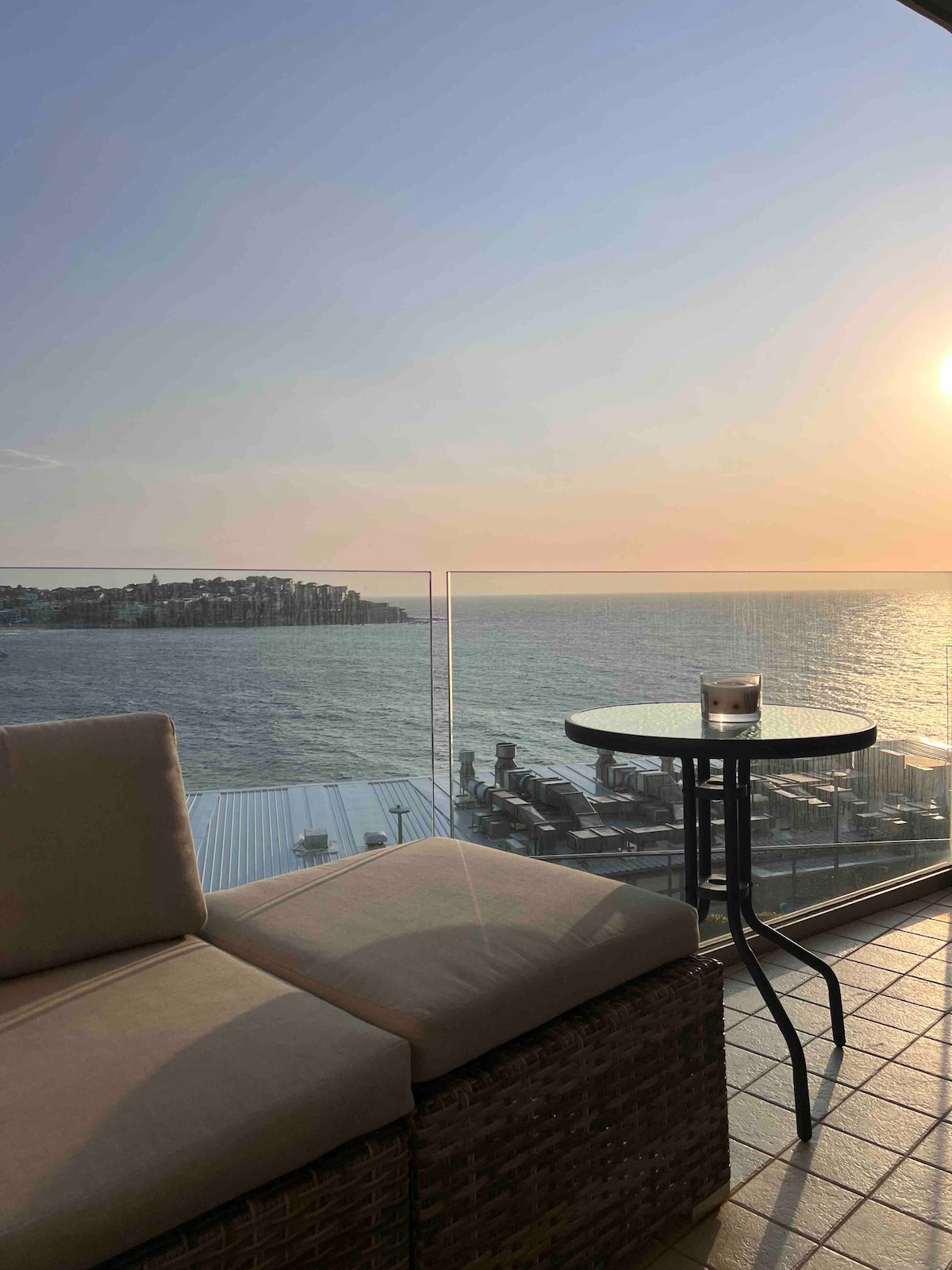 A corner seating area is visible, featuring a plush cushion against a glass balcony railing. A small round table is positioned nearby, holding a drink. The ocean stretches into the distance, reflecting soft hues of sunset, enhancing the serene atmosphere.