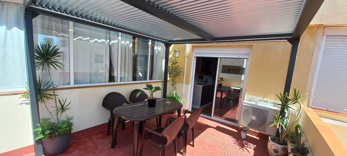 A covered terrace is featured, furnished with a table and four chairs. Potted plants are arranged around the space, providing a natural touch. Large windows bathe the area in light, while an air conditioning unit is visible, ensuring comfort during warmer days.