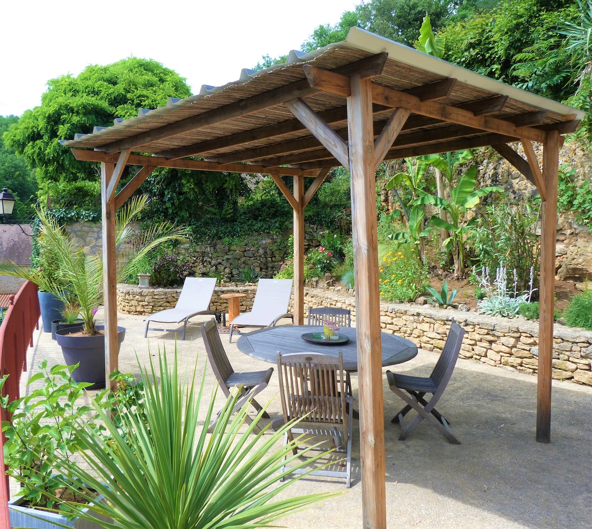 A shaded outdoor terrace features a wooden gazebo supported by sturdy posts, surrounded by lush greenery. A circular wooden table is accompanied by several chairs, while two lounge chairs are positioned nearby. Potted plants add to the inviting ambiance of the space.