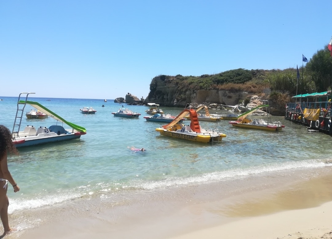 A sandy beach is depicted with clear blue waters and a variety of boats anchored nearby. The colorful rental boats, featuring yellow, red, and white shades, are easily identifiable. Lush greenery lines the shore in the background, contributing to a serene coastal setting.